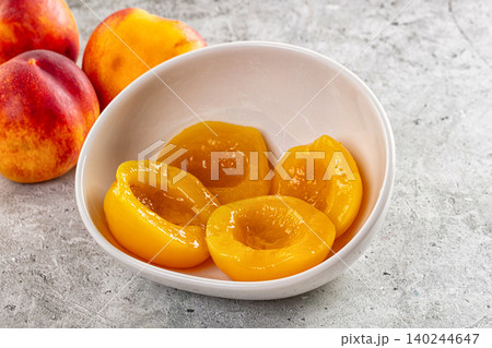 canned peach halves in white bowl with fresh peaches on stone surface 140244647