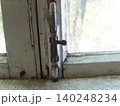 Captures a close-up of an old, weathered wooden window frame with peeling white paint. A vintage metal bolt lock secures the sash, while bright natural light streams from the outside 140248234