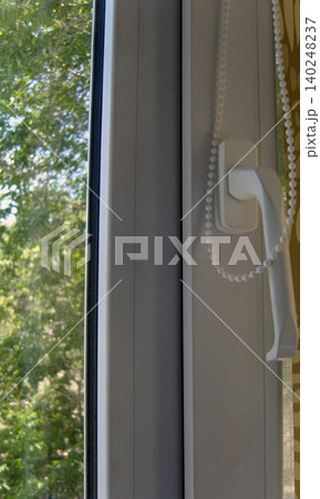 Modern white uPVC window. The focus is on the frame, the ergonomic handle, and the beaded pull chain for a pull-down blind. The handle is positioned for right-hand use Modern white uPVC window. The focus is on the frame, the ergonomic handle, and the beaded pull chain for a pull-down blind. The handle is positioned for right-hand use 140248237