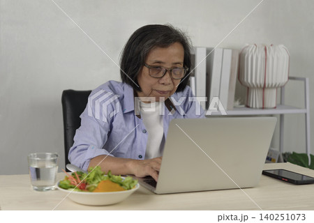 Senior Asian woman working on laptop and typing while eating healthy salad lunch at home. Elderly digital lifestyle, remote work, freelance, and multitasking concept. Senior Asian woman working on laptop and typing while eating healthy salad lunch at home. Elderly digital lifestyle, remote work, freelance, and multitasking concept. 140251073