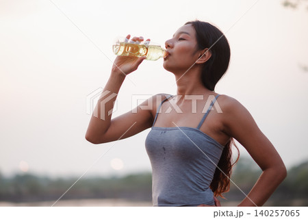 Young woman drinking water after outdoor yoga by a calm lakeside at sunrise, showing hydration, wellness, recovery, fitness, refreshment, healthy lifestyle, and active self-care in nature. 140257065