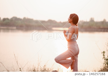 Young woman practicing tree pose yoga by a tranquil lakeside at golden sunset, showing balance, wellness, mindfulness, flexibility, healthy lifestyle, nature fitness, and calm energy. Young woman practicing tree pose yoga by a tranquil lakeside at golden sunset, showing balance, wellness, mindfulness, flexibility, healthy lifestyle, nature fitness, and calm energy. 140257168