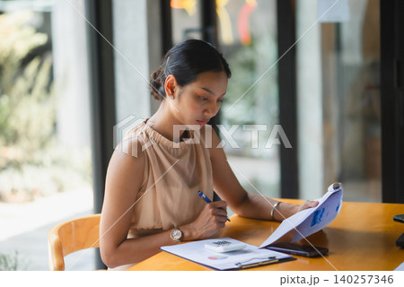 Businesswoman writing notes while reviewing financial report, calculator, and smartphone at modern office table, showing accounting, data analysis, budgeting, planning, and focused work. Businesswoman writing notes while reviewing financial report, calculator, and smartphone at modern office table, showing accounting, data analysis, budgeting, planning, and focused work. 140257346