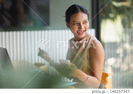 Cheerful Asian businesswoman holding smartphone beside laptop in modern cafe office, smiling during client communication, financial planning, online work, accounting, productivity, and success. Cheerful Asian businesswoman holding smartphone beside laptop in modern cafe office, smiling during client communication, financial planning, online work, accounting, productivity, and success. 140257347