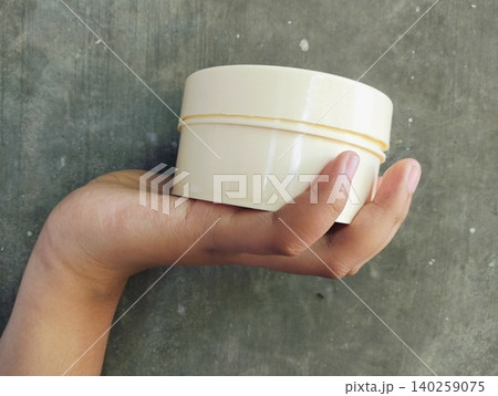 Hand Holding White Cosmetic Cream Jar on Concrete Wall Background 140259075