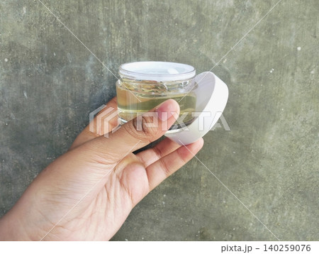 Hand Holding Transparent Cosmetic Gel Jar on Concrete Wall Background Hand Holding Transparent Cosmetic Gel Jar on Concrete Wall Background 140259076