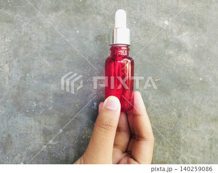 Hands Holding Cosmetic Serum Dropper and Red Glass Bottle on Concrete Background Hands Holding Cosmetic Serum Dropper and Red Glass Bottle on Concrete Background 140259086