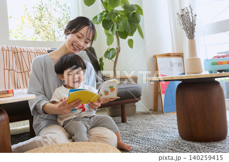 リビングで母親の膝に座り絵本を読む笑顔の幼児 リビングで母親の膝に座り絵本を読む笑顔の幼児 140259415
