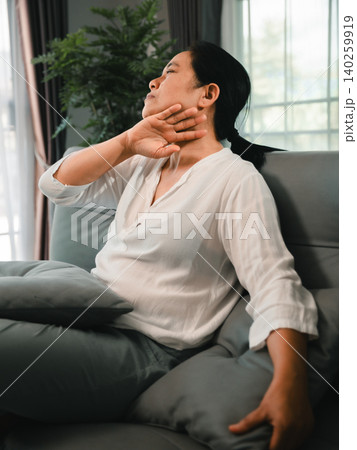 Asian woman sitting on a sofa at home with neck discomfort, stretching her shoulder and throat area, representing muscle tension, fatigue, stress relief, wellness, and body care routine. Asian woman sitting on a sofa at home with neck discomfort, stretching her shoulder and throat area, representing muscle tension, fatigue, stress relief, wellness, and body care routine. 140259919