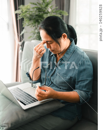 Tired Asian woman using a laptop at home, holding glasses and touching her nose, showing eye strain, screen fatigue, remote work stress, headache, digital burnout, and online productivity. Tired Asian woman using a laptop at home, holding glasses and touching her nose, showing eye strain, screen fatigue, remote work stress, headache, digital burnout, and online productivity. 140259928