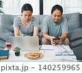 Asian family sitting together on a sofa with a laptop, smartphone, documents, and toast at home, showing remote work, budgeting, teamwork, online tasks, and daily family life routine. 140259965