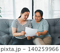 Asian parent and young woman reading a document together on a sofa at home, showing family planning, paperwork, financial review, support, trust, advice, household decisions, and care. 140259966