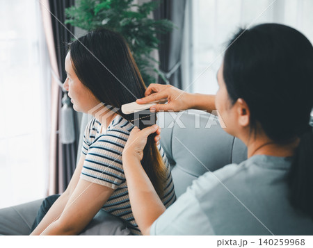 Asian parent gently brushing a young woman's long hair on a sofa at home, showing family bonding, hair care, caregiving, beauty routine, tenderness, support, comfort, and daily love. 140259968