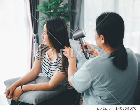 Asian parent drying a young woman's long hair with a hair dryer on a sofa at home, showing family care, beauty routine, grooming, support, bonding, everyday care, and domestic lifestyle. 140259970