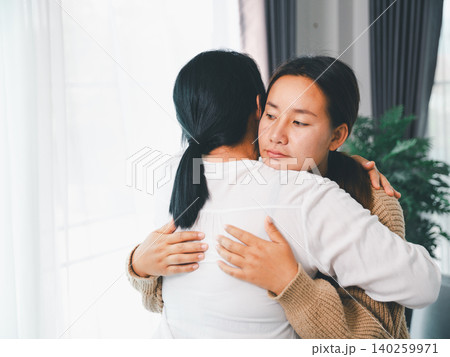 Asian parent hugging a young woman at home, showing family comfort, emotional support, empathy, reconciliation, mental wellness, caregiving, trust, and a healing relationship in a bright living room. 140259971