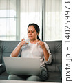 Asian woman using a laptop on a sofa at home while drinking water, showing remote work, online meeting, healthy break, home office lifestyle, hydration, and daily productivity routine. 140259973