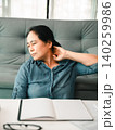 Asian woman with neck pain sitting at a home desk, stretching beside an open notebook and glasses, showing office fatigue, poor posture, body tension, wellness, and work stress relief. 140259986