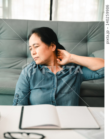 Asian woman with neck pain sitting at a home desk, stretching beside an open notebook and glasses, showing office fatigue, poor posture, body tension, wellness, and work stress relief. Asian woman with neck pain sitting at a home desk, stretching beside an open notebook and glasses, showing office fatigue, poor posture, body tension, wellness, and work stress relief. 140259986