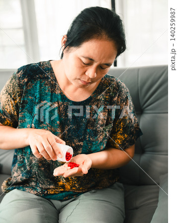 Asian woman pouring red pills from a bottle into her hand while sitting on a sofa, representing medication routine, healthcare, treatment, wellness, home care, and personal health. Asian woman pouring red pills from a bottle into her hand while sitting on a sofa, representing medication routine, healthcare, treatment, wellness, home care, and personal health. 140259987