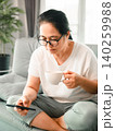Asian woman wearing glasses holding a coffee cup and smartphone on a sofa at home, showing mobile browsing, morning routine, online communication, lifestyle comfort, and relaxation. 140259988