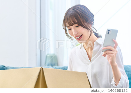 届いた荷物を前にスマホを見て驚く女性 届いた荷物を前にスマホを見て驚く女性 140260122