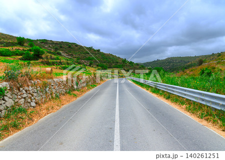 Scenic country road winds through rolling agricultural hills under a cloudy sky Scenic country road winds through rolling agricultural hills under a cloudy sky 140261251