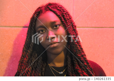 Night portrait of a confident young Black woman illuminated by pink neon lights against an urban wall Night portrait of a confident young Black woman illuminated by pink neon lights against an urban wall 140261335