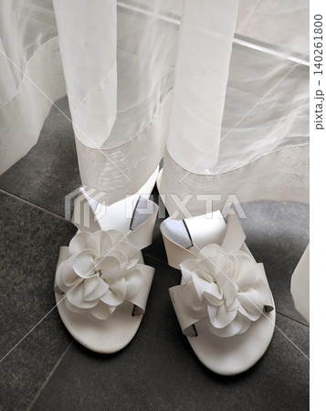 A pair of elegant white slides, adorned with voluminous flowers, rests beside a semi-transparent curtain A pair of elegant white slides, adorned with voluminous flowers, rests beside a semi-transparent curtain 140261800