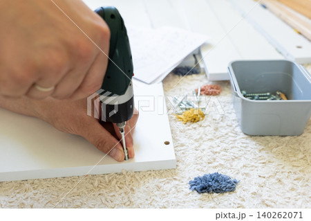 Hand using electric screwdriver to assemble furniture on carpeted floor with screws and tools nearby Hand using electric screwdriver to assemble furniture on carpeted floor with screws and tools nearby 140262071