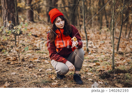 Woman crouches in autumn forest while holding cluster of wild mushrooms. Mushroom hunting tips, forest biology exploration, field guide reference, learning about fungi. 140262193