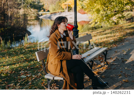 Woman sits on park bench drinking from reusable mug near water during autumn. Slow living autumn routine, digital detox nature getaway, cozy fall seasonal lifestyle. Woman sits on park bench drinking from reusable mug near water during autumn. Slow living autumn routine, digital detox nature getaway, cozy fall seasonal lifestyle. 140262231
