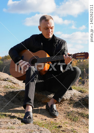 Portrait of smiling caucasian artist plays wooden acoustic guitar in autumnal park. Young songwriter plays string musical instrument hobby outside in nature fall time. Audio music healing 140262311