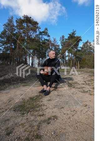 Blonde man playing string guitar outdoors in autumn forest. Concept of sound therapy, mental health and wellness rituals. Calmness tranquility audio-sensory practices. Aura farming energy 140262319