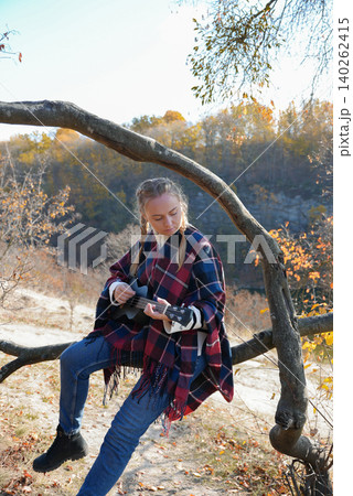 Beautiful blonde girl playing guitar ukulele and singing relaxing enjoying outdoors on sunny autumn day. Hippie woman in boho style training playing musical instrument. Fall leaves Beautiful blonde girl playing guitar ukulele and singing relaxing enjoying outdoors on sunny autumn day. Hippie woman in boho style training playing musical instrument. Fall leaves 140262415