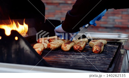 Street food chef wearing blue gloves grilling sausages over flaming barbecue, carefully monitoring cooking process for optimal flavor Street food chef wearing blue gloves grilling sausages over flaming barbecue, carefully monitoring cooking process for optimal flavor 140262535