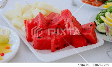 Vibrant watermelon and melon slices decorating white rectangular plate, surrounded by colorful buffet selection of fresh fruits and cheeses 140262769