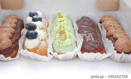 Assorted pastel eclairs arranged in rows on paper trays, variety includes blueberry, pistachio and chocolate flavors, bright market setting, inviting textures, suitable for catering catalog and menu 140262788