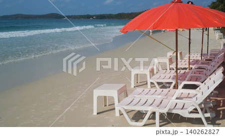White empty beach lounge chairs under a red beach umbrella on clean sandy beach with calm sea in background. Beach relaxation concept White empty beach lounge chairs under a red beach umbrella on clean sandy beach with calm sea in background. Beach relaxation concept 140262827