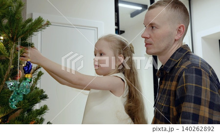 Young girl hanging delicate ornaments, father watching with warm smile, creating tender holiday moment beside glowing christmas tree Young girl hanging delicate ornaments, father watching with warm smile, creating tender holiday moment beside glowing christmas tree 140262892