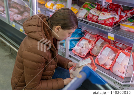 Woman reading frozen chicken label closely, checking ingredients and expiry while holding cart handle at refrigerated display, focused expression, prepping weeknight dinner and comparing value Woman reading frozen chicken label closely, checking ingredients and expiry while holding cart handle at refrigerated display, focused expression, prepping weeknight dinner and comparing value 140263036