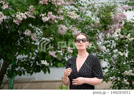 Woman in black polka dot dress poses beside blooming lilac, with spring flowers and historic architecture creating refined cultural atmosphere. Cultural tourism, lifestyle, heritage, urban travel Woman in black polka dot dress poses beside blooming lilac, with spring flowers and historic architecture creating refined cultural atmosphere. Cultural tourism, lifestyle, heritage, urban travel 140263051