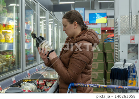 Woman inspecting frozen dumplings in supermarket freezer, comparing ingredients and nutrition for quick weekday dinner brown jacket, shopping cart, focused expression, fluorescent lighting, product Woman inspecting frozen dumplings in supermarket freezer, comparing ingredients and nutrition for quick weekday dinner brown jacket, shopping cart, focused expression, fluorescent lighting, product 140263059