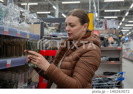 Caucasian Woman Inspecting Glass Vase In Store Aisle, Wearing Brown Puffer Jacket, Holding Green Glass, Shopping Cart Visible, Fluorescent Lighting, Focused Expression, Comparing Textures And Price. Caucasian Woman Inspecting Glass Vase In Store Aisle, Wearing Brown Puffer Jacket, Holding Green Glass, Shopping Cart Visible, Fluorescent Lighting, Focused Expression, Comparing Textures And Price. 140263072