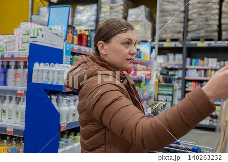 Woman Selecting Shower Sponge In Store, Focused Expression Scanning Texture And Label, Brown Puffer Jacket, Packed Shelves Of Toiletries, Fluorescent Lighting, Everyday Errand Mood. 140263232