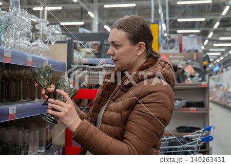 Woman Compares Prices. Woman In Puffer Coat Examines Product Price And Label Details Diligently. Female Shopper Wearing Padded Jacket Inspects Price Tags And Discount Information Attentively Woman Compares Prices. Woman In Puffer Coat Examines Product Price And Label Details Diligently. Female Shopper Wearing Padded Jacket Inspects Price Tags And Discount Information Attentively 140263431