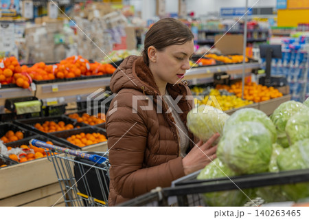 Woman Examining Cabbage For Quality In Supermarket, Brown Coat, Checking Leaves And Texture, Produce Bins And Bright Signage Visible, Planning Meals And Checking Freshness Before Purchase Woman Examining Cabbage For Quality In Supermarket, Brown Coat, Checking Leaves And Texture, Produce Bins And Bright Signage Visible, Planning Meals And Checking Freshness Before Purchase 140263465