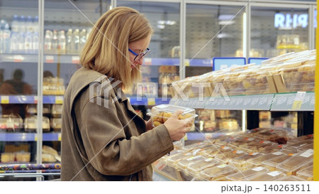 Young woman grocery shopping, choosing cookies in supermarket, reading label and holding plastic box with dessert, standing in front of shelves full of various products Young woman grocery shopping, choosing cookies in supermarket, reading label and holding plastic box with dessert, standing in front of shelves full of various products 140263511
