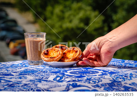Portuguese custard tart and hand serving pastries beside coffee on a blue table in sunny outdoor light. Food tourism, culinary travel, local cuisine, traditional dessert 140263535
