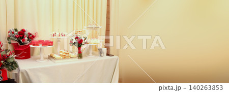 Festive candy bar with sweets, cakes and red rose bouquets on a white table. Wide panoramic banner of wedding or birthday dessert buffet with a large copy space on a cream wall background Festive candy bar with sweets, cakes and red rose bouquets on a white table. Wide panoramic banner of wedding or birthday dessert buffet with a large copy space on a cream wall background 140263853
