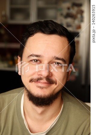 Portrait of a smiling young man with a beard and mustache looking at camera. Close-up headshot of a happy confident guy indoors with a blurred cozy background Portrait of a smiling young man with a beard and mustache looking at camera. Close-up headshot of a happy confident guy indoors with a blurred cozy background 140263915